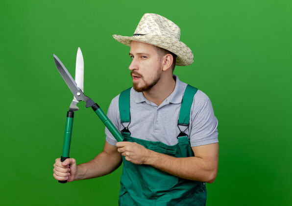 帽子自信的年轻英俊的斯拉夫园丁 穿着制服 戴着帽子 手里拿着修剪器男人花园绿色