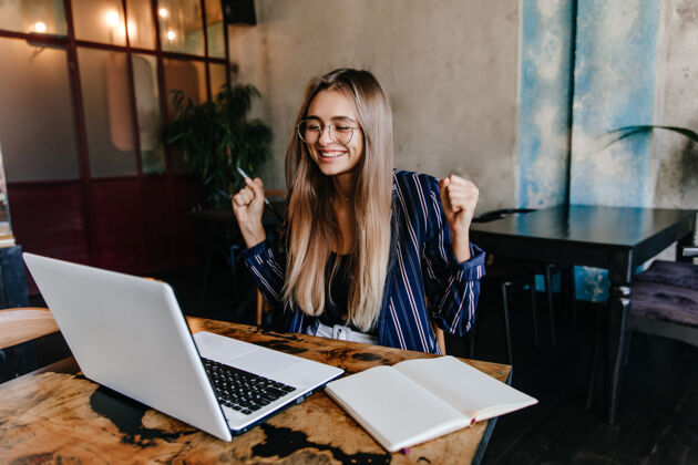 快乐兴奋的长发女孩在与电脑的工作乐趣微笑的女自由职业者在舒适的咖啡馆使用笔记本电脑的室内照片成人年轻手机