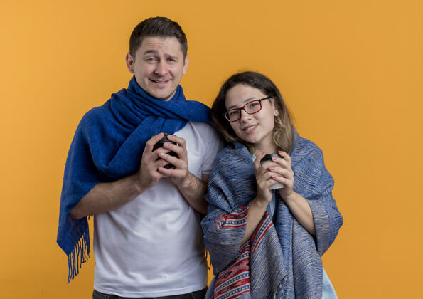 杯子冻得幸福的年轻夫妇一男一女裹着温暖的毯子 手里拿着咖啡杯 脸上挂着橙色的微笑温暖冷冻咖啡