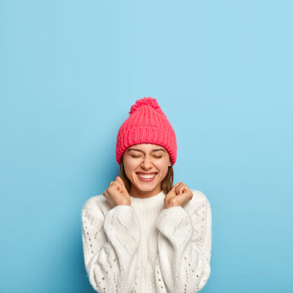 时尚快乐的小女孩喜出望外 握紧拳头 心情很好 穿着白毛衣 戴着粉红色的帽子 在寒冷的秋日里穿着暖和的衣服 孤零零地站在蓝色的墙上自然舒适编织