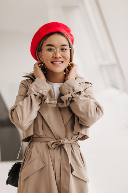 休息微笑的棕色眼睛的亚洲女人的肖像 戴着时髦的眼镜 法式红色贝雷帽和米色风衣 靠着窗户看前面年轻卷发亚洲人