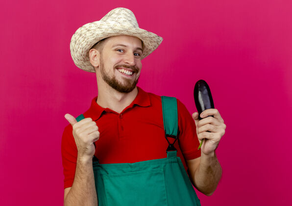 大拇指面带微笑的年轻英俊的斯拉夫园丁 穿着制服 戴着帽子 手里拿着茄子 抬起大拇指制服年轻微笑