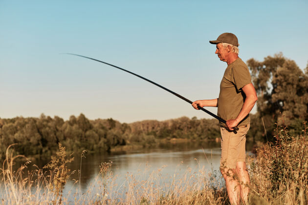 运动渔夫站在湖畔或河畔望着手中的鱼竿 在夕阳下垂钓 在美丽的大自然中 穿着绿色的t恤和裤子湖休闲海岸