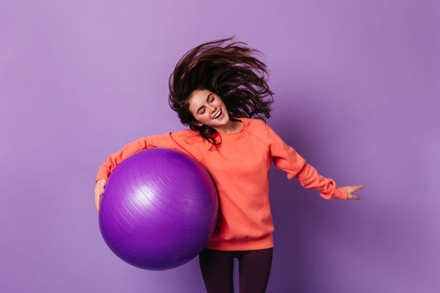 黑发身穿橙色运动衫的黑发女子把玩着头发 在隔离墙上拿着紫色的健身球头发健美操瑜伽垫