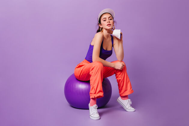 卷发戴着帽子和运动紫橙色套装的女人坐在fitball上 手里拿着牛奶巧克力身材锻炼皮肤