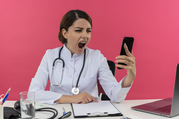 医疗愤怒的年轻女医生穿着医用长袍和听诊器坐在办公桌旁 手里拿着医疗工具和笔记本电脑 手里拿着手机 看着手机专业女士穿