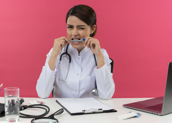长袍年轻的女医生穿着医用长袍和听诊器坐在办公桌旁 手里拿着医疗工具和笔记本电脑 眨眼着 咬着笔眨眼听诊器专业