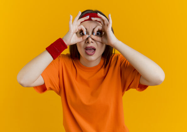 橙色令人印象深刻的年轻运动女子戴着头带和腕带做看手势看用手作为望远镜佩戴手势年轻