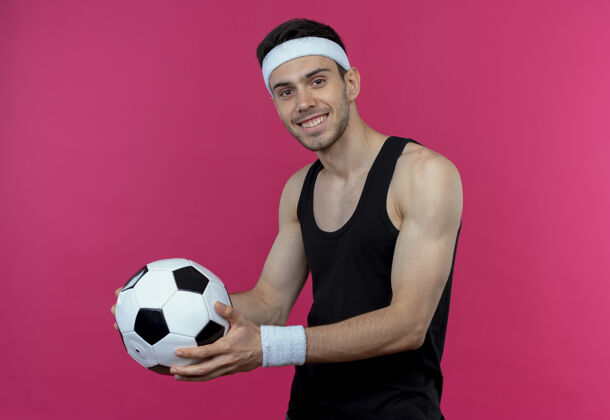 微笑戴着头巾 手持足球 站在粉红色的墙上欢快微笑的年轻人看抱着站着