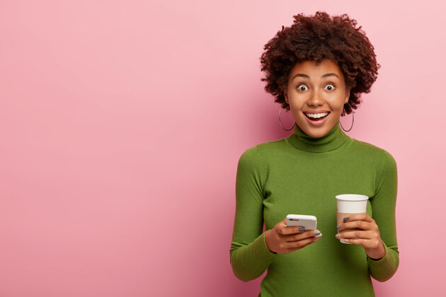 移动快乐的卷发女人 健康的皮肤 穿着休闲的绿色套头衫 喝着现代的手机和外卖咖啡 享受在线交流站着社交信息