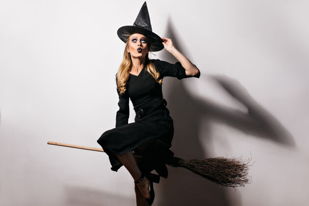 神秘身着黑色长裙 头戴魔法帽 准备参加狂欢节的快乐女模特室内拍摄的金发女巫拿着旧扫帚传统金发女人