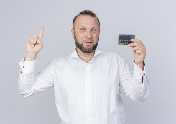 男人胡子男人穿着白衬衫展示信用卡展示食指笑脸有新想法站在白墙上穿目录站