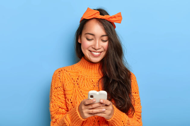 年轻时尚的黑发千禧一代女孩忙着查看她的邮箱 拿着手机 头上系着橙色的头带 穿着暖和的毛衣套头衫人微笑