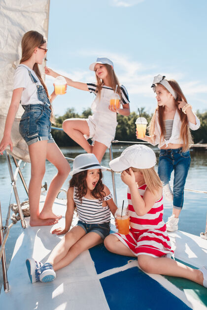 衣服孩子们在游艇上喝着橙汁青少年或少女们在蓝天下户外五颜六色的衣服孩子们的时尚 阳光明媚的夏天 河流和假日的概念水手朋友旅游