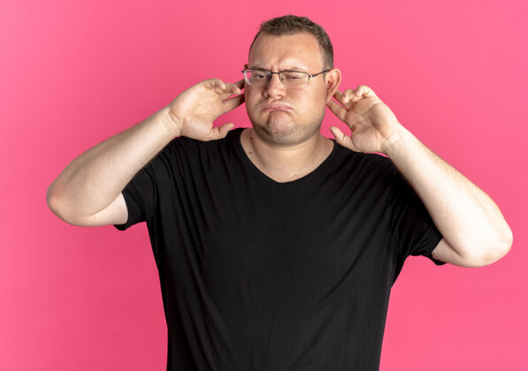 表情一个戴眼镜 身穿黑色t恤的超重男子站在粉红色的墙上 用手指捂住耳朵 脸上带着恼怒的表情戴着手指超重