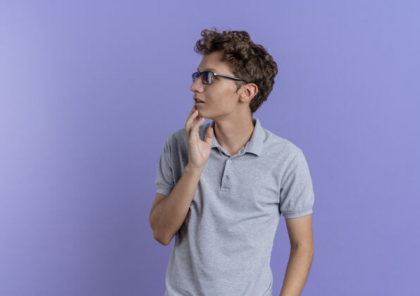 马球一个戴着黑眼镜 穿着灰色马球衫的年轻人站在蓝色的墙上 茫然地看着一边帅哥穿站