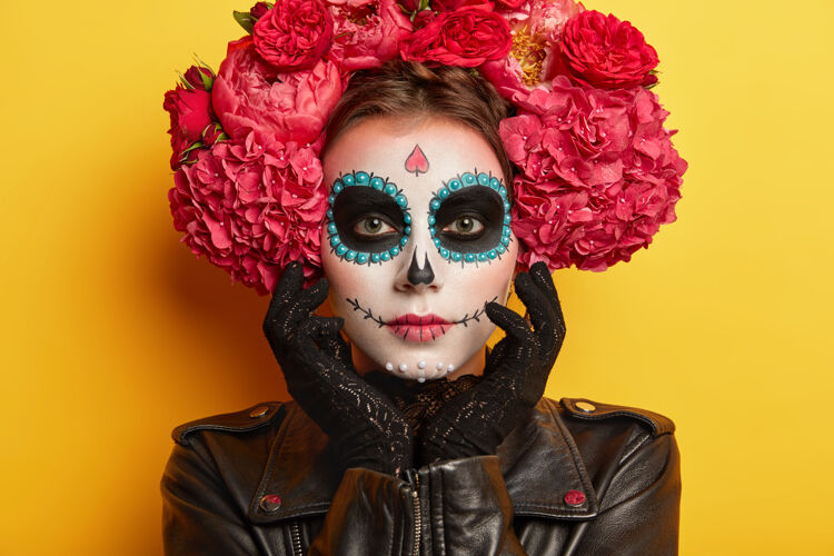 皮革头像的严肃美丽的女人头像与头骨化妆 画的脸由艺术家 穿黑色衣服 想看起来幽灵 在黄色的背景下摆姿势传统的墨西哥节日室内微笑头骨