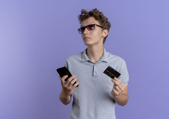 男人一个戴着黑眼镜 穿着灰色马球衫 拿着智能手机和信用卡的年轻人站在蓝色的墙上困惑地看着我穿拼图手机