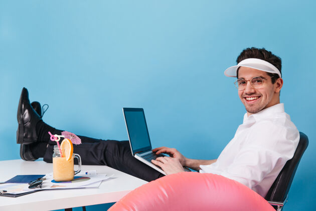 正式戴着眼镜和帽子的男人在一个孤立的空间里工作男人拿着笔记本电脑 鸡尾酒和充气圈摆姿势文件成功员工
