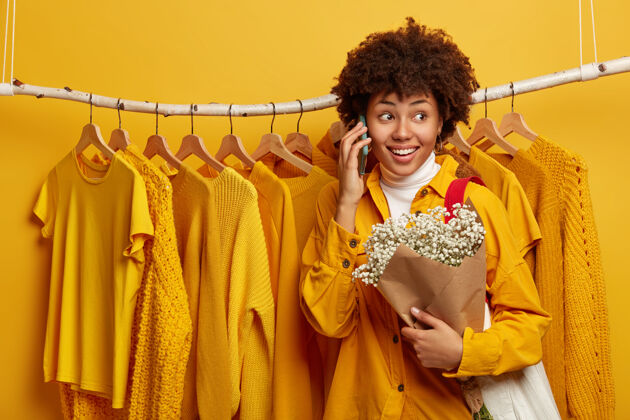 买家快乐的卷发女人 带着快乐的表情 打电话给朋友 捧着漂亮的花束 拎着包 对着破烂的黄亮衣服摆姿势时尚服装造型师