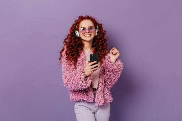 羊毛快乐的红发女孩穿着粉色环保外套和浅色裤子微笑着戴着蓝色耳机的女人拿着黑色智能手机上衣少女魅力
