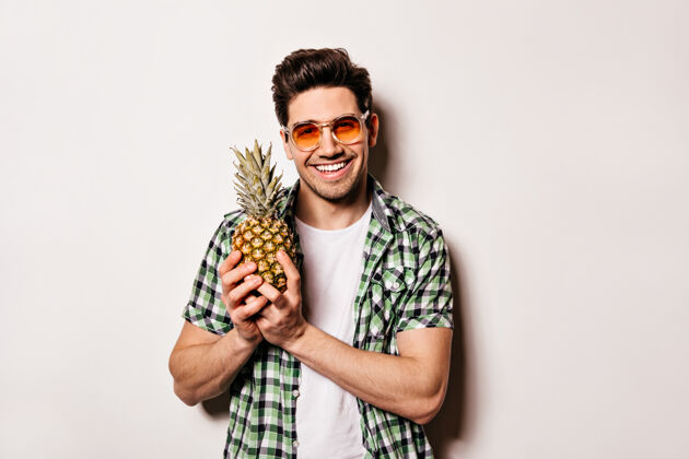 休息一个戴着橘色眼镜 穿着格子绿衬衫的黑发男人微笑着 手里拿着菠萝在白色的空间里孤独男性水果