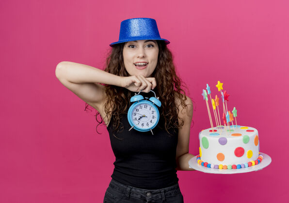 抱着一位年轻漂亮的女士 卷发 戴着节日礼帽 手里拿着生日蛋糕和闹钟 站在粉色的墙上 看起来很惊讶 生日快乐卷发女人年轻