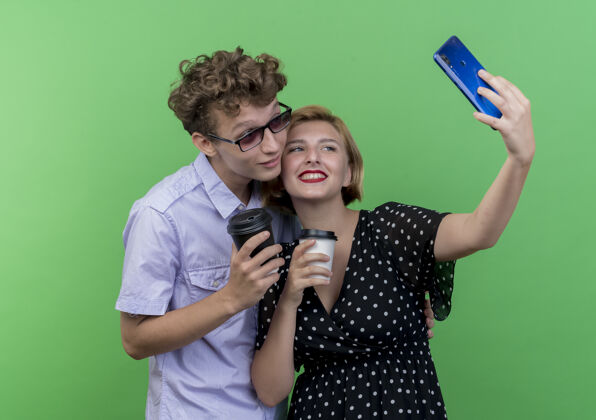 用着年轻漂亮的夫妇站在一起拿着咖啡杯用手机自拍微笑着越过绿色的墙壁微笑手机抱着