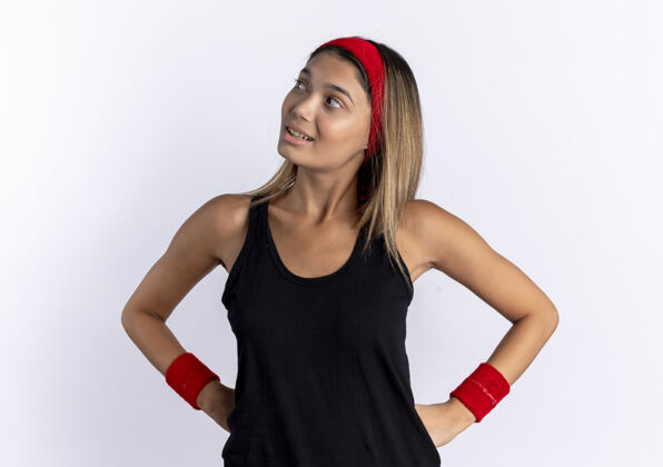 年轻身穿黑色运动服 头戴红色头巾的年轻健身女孩站在白墙上 自信地微笑着 双臂放在臀部自信运动装臀部