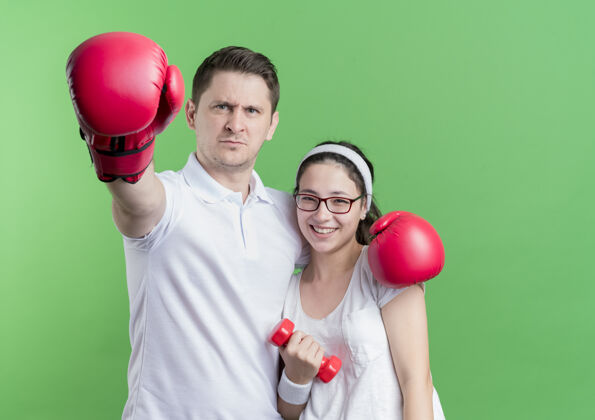 年轻一对年轻的运动情侣 一个手持哑铃的女人和一个戴拳击手套的男人微笑着站在绿色的墙上哑铃男人运动