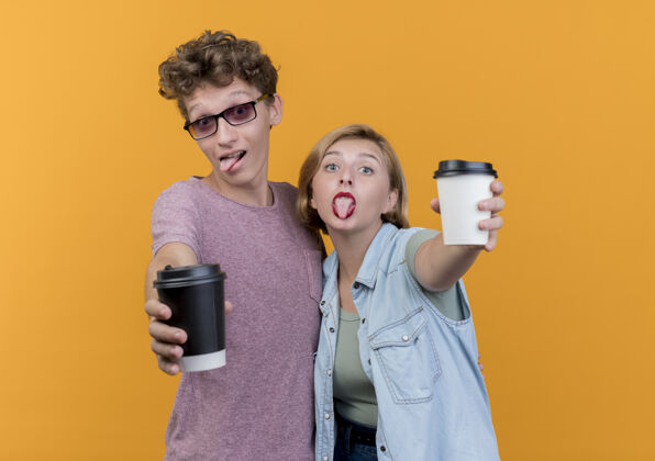 情侣一对年轻漂亮的情侣穿着休闲服展示咖啡杯摆姿势 微笑着伸出舌头站在橙色的墙上表情女人坚持