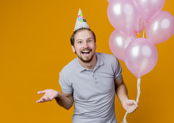 帽子戴着节日帽的年轻人举着气球欢笑着站在橙色的墙上庆祝生日抱着气球男人