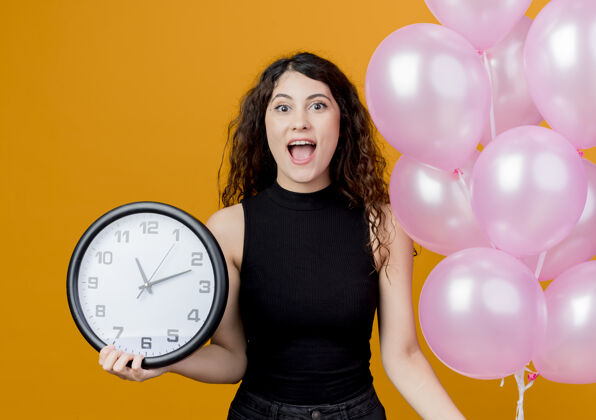 气球年轻漂亮的女人 卷发 手持一束气球和挂钟 站在橙色的墙上 快乐而兴奋的生日派对年轻墙壁站着