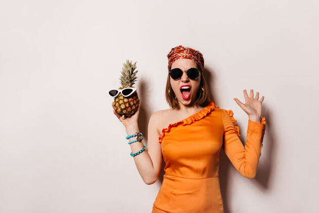 菠萝情绪化的女人戴着时髦的头带 穿着橙色的裙子 自命不凡地尖叫着 戴着太阳镜拿着菠萝情绪手生命