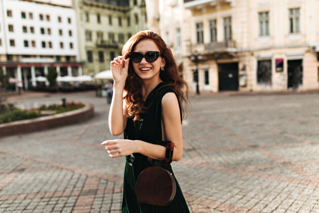 姿势戴着太阳镜的时尚女士在城市中穿行头发连衣裙红色头发