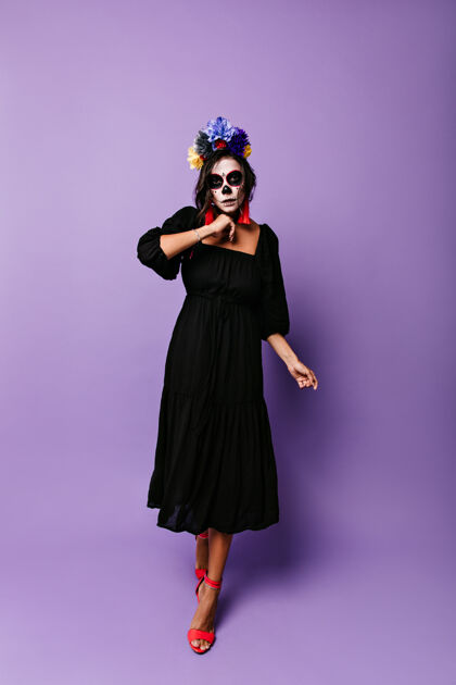 恶魔穿黑色迷笛裙的女孩靠着紫色的墙走着戴着头盖骨面具的模特摆出万圣节的姿势嘉年华花死亡之日