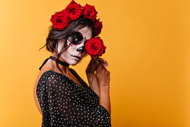 化妆穿着万圣节服装的迷人成熟女人喜欢玫瑰墨西哥女人用红花闭上眼睛的特写肖像年轻恶魔墨西哥