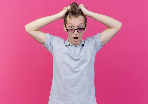 站一个戴着黑色眼镜 穿着灰色马球衫的沮丧的年轻人 站在粉红色的墙上 摸着头 拉着头发穿拉年轻