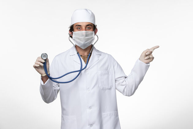 冠状病毒预防正面图：男医生 穿着医疗服 戴着口罩 以防柯维德-在白色办公桌上使用听诊器-柯维德-大流行办公桌医疗穿着