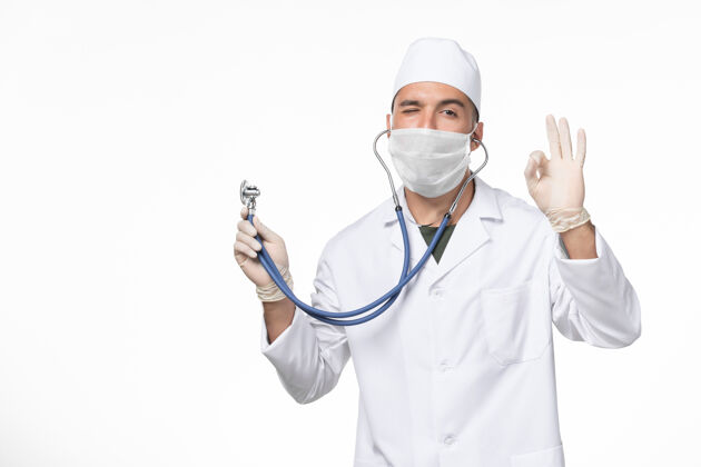 听诊器正面图男医生穿着医疗服 戴着口罩因冠状病毒-用听诊器眨眼白墙冠状病毒-大流行病毒病实验室外套口罩男性