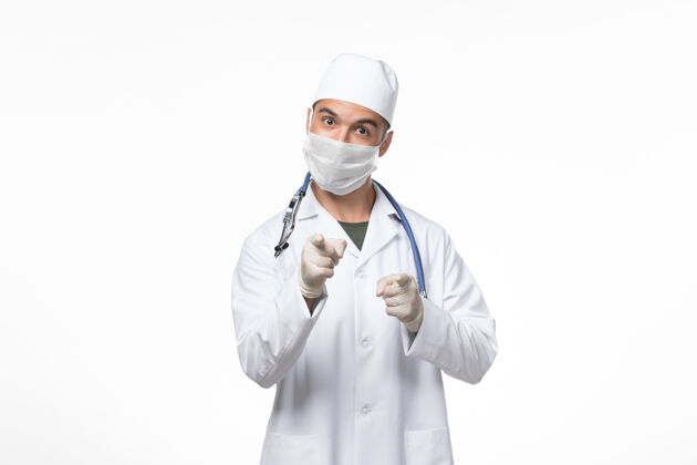 听诊器正面图：男医生穿着医疗服 戴着防冠状病毒口罩 用蓝色听诊器对着白墙上的冠状病毒病大流行病毒口罩大流行冠状病毒
