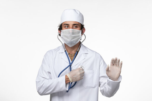 冠状病毒防护正面图：男医生穿着医疗服 戴着口罩以防病毒感染-使用听诊器对白墙病病毒进行病毒感染-大流行视图病毒使用