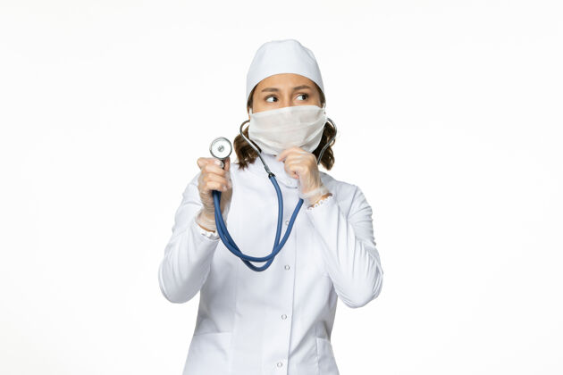 视图前视图穿着白色医疗服和口罩的女医生在使用听诊器和思考白墙潘多米病毒疾病的药物正面病毒套装