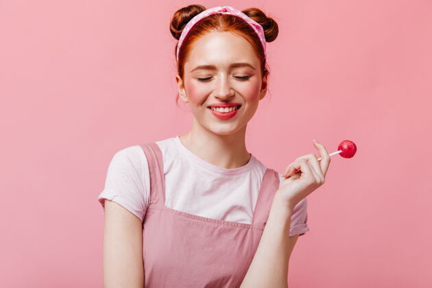 优雅穿着白色t恤和上衣的快乐女人微笑着拿着粉色背景的糖果红发糖果粉色背景