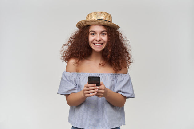 女士有魅力的成年红头发女孩的肖像 卷发穿着条纹露肩上衣和帽子拿着智能手机和微笑隔着白墙孤立快乐室内外观