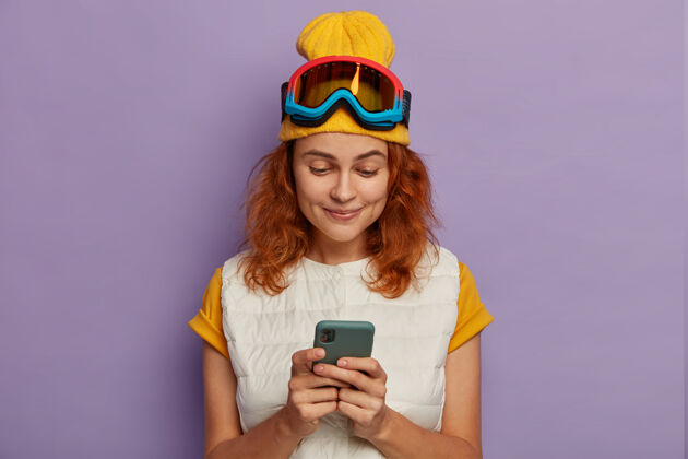 护目镜活跃的红发少女照片使用手机上网聊天 在山上度过寒假 戴滑雪板防护面具 享受旅游和免费上网情绪白种人冬季