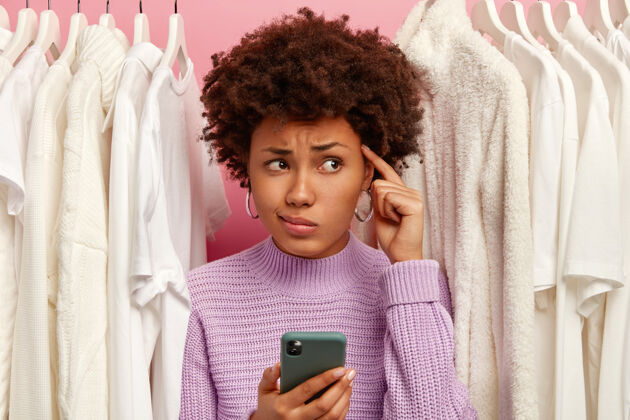 买家不幸的黑皮肤女人透过衣柜看 站在衣架上的白色衣服之间 食指放在太阳穴上 手里拿着现代手机女士毛衣移动电话