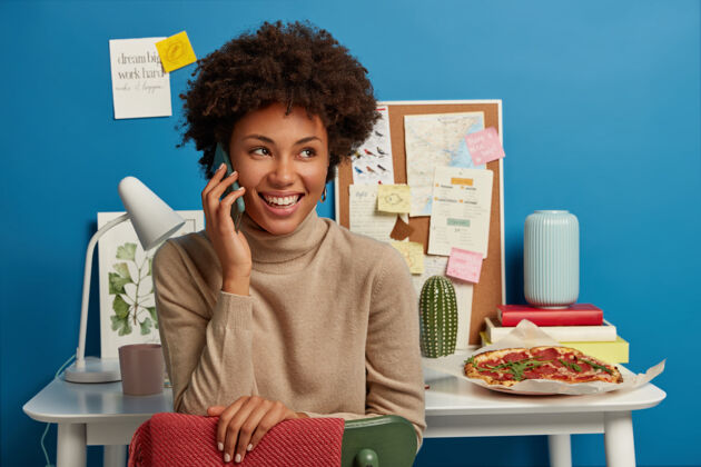 高兴高兴的黑皮肤女人有电话交谈 看向别处 心情很好完成工作 坐在桌上与一堆笔记本 墙板上的便签 美味的比萨饼比萨饼享受女士