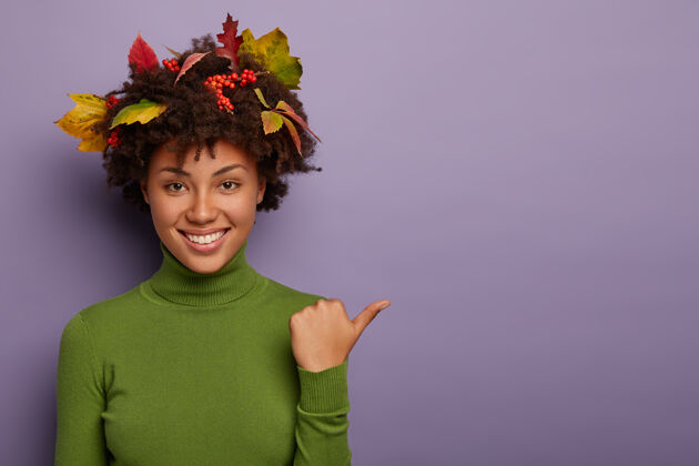 指示器快乐的女人 卷发被秋天的树叶装饰着 在室内摆姿势 拇指放在一边 穿着绿色休闲套头衫 紫色背景下孤立 笑容灿烂卷发肖像欢呼
