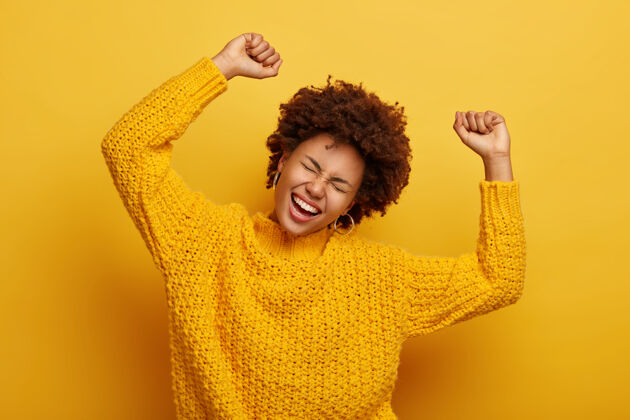 空白快乐的非洲女人举起双臂 歪着头 穿着休闲的针织套头衫 从快乐中欢笑 庆祝胜利 孤立在黄色上复制空间喜悦针织